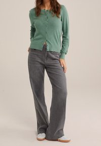 Woman wearing a textured green buttoned cardigan, high-waisted wide-leg gray jeans, and white sneakers on a neutral background.