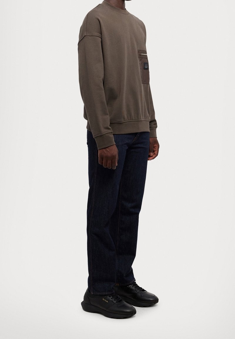 Homme portant un sweat-shirt marron à manches longues avec poche avant, un jean bleu foncé et des baskets noires décontractées, debout devant un fond uni.