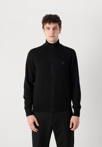 Svart zip-up tröja med hög krage, tillverkad av slätt stickat material. Har en liten fyrkantig logotyp på vänster bröst och ribbade muddar.