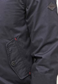 Mörkblå vattentät jacka i slät tyg, med en ficka som stängs med knapp och en märkespatch på axeln. Röda sömmar synliga.