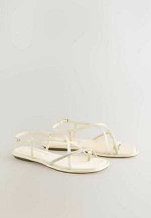 Valkoiset tasapohjaiset sandaalit, joissa on ohuet hihnat ja pieni kimalteleva yksityiskohta varpaiden ylle. Pehmeä, teksturoitu pohja ja säädettävä nilkkahihna.
