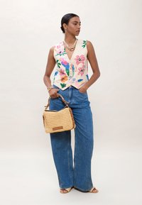 Vrouw draagt een mouwloos vest met bloemenborduursels, wijde blauwe jeans, houdt een geweven handtas vast, gecombineerd met gouden sieraden en naakte sandalen.