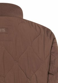 Brązowa pikowana kurtka z przeszyciami w kształcie fali i prążkowanym kołnierzem, widoczna górna część pleców i ramiona.