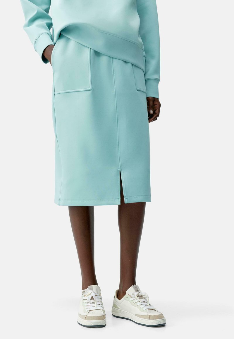 Menta zöld midi szoknya sima anyagból, elöl hasítékkal, egy nagy zsebbel kiemelve, fehér és krémszínű sportcipőkkel párosítva.
