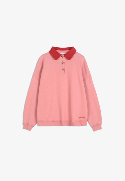 Růžový pulovr s dlouhými rukávy a červeným límečkem, tři knoflíky u krku, žebrované manžety a malé červené logo blízko lemu na bílém pozadí.