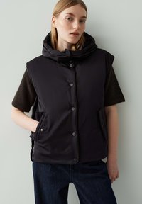 Gepolsterte schwarze Weste mit hohem Kragen und Druckknopfverschluss, ausgestattet mit Seitentaschen und kurzen Ärmeln, kombiniert mit dunklen Jeans.