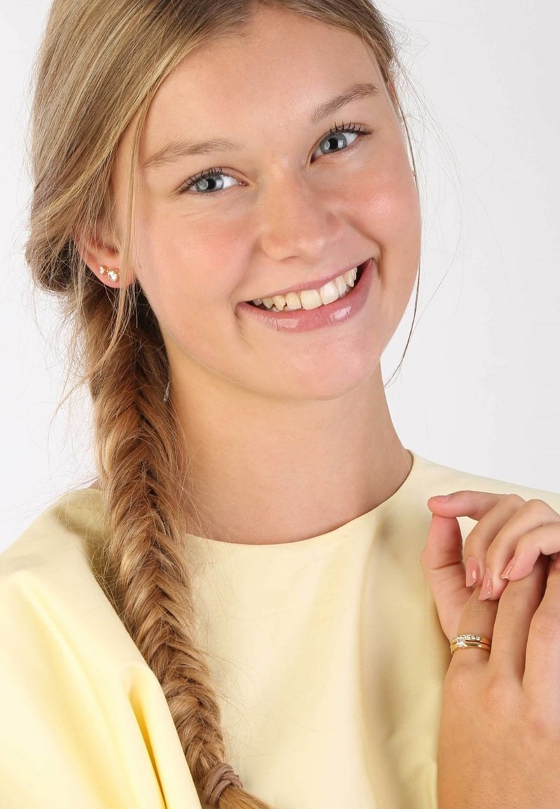 Een jonge persoon met een gevlochten kapsel draagt een gele top, minimale oorbellen en een gouden ring met een heldere steendetail.