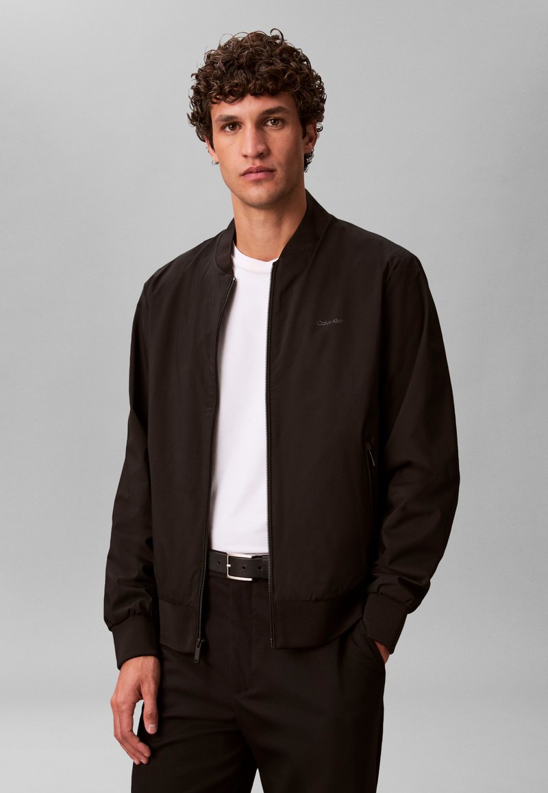 Jeune homme aux cheveux bouclés portant une veste noire zippée, un t-shirt blanc, une ceinture noire et un pantalon noir, debout devant un fond gris.