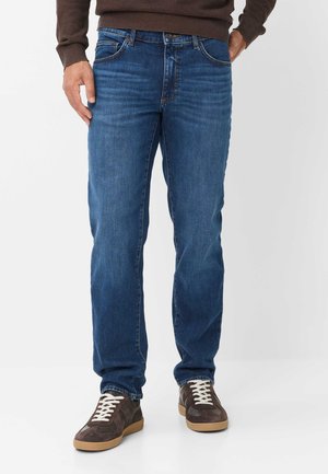 Mann trägt blaue Straight-Leg-Jeans, braunen Pullover und dunkelbraune Sneakers mit weißen Schnürsenkeln, steht vor weißem Hintergrund.