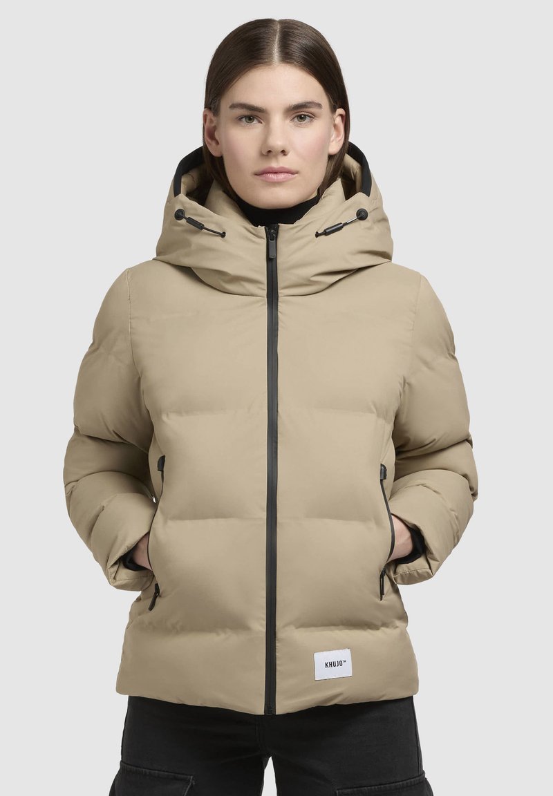 Beige Steppjacke mit hohem Kragen, schwarzen Reißverschlüssen und verstellbare Kapuze. Verfügt über Seitentaschen und einen Logo-Patch auf der unteren Vorderseite.
