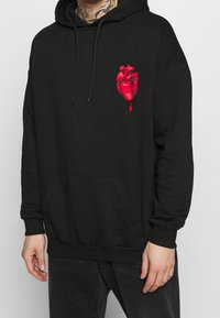 Persona con sudadera negra con un gráfico de un corazón anatómico rojo y una gota de sangre en el lado izquierdo del pecho, combinada con pantalones negros.