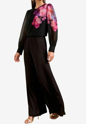 Kobieta w czarnym kombinezonie z przezroczystymi długimi rękawami i różowym kwiatowym wzorem na górnej części klatki piersiowej i rękawach, zestawiona ze złotymi sandałami na otwartym palcu.