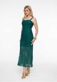 Türkisfarbenes Spitzenkleid mit angepasstem Oberteil und spaghetti-Trägern; durchsichtiger Rock mit gewelltem Saum, kombiniert mit goldenen Plateausandalen und Accessoires.