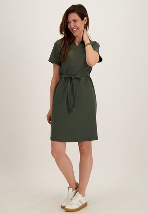 Vrouw die glimlacht, draagt een olijfgroene korte mouw jurk met een strik in de taille en witte sneakers, staand tegen een effen lichte achtergrond.