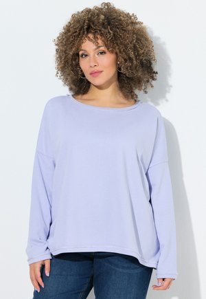 Kvinde med krøllet hår iført en lyse lavendel langærmet top og mørkeblå jeans, stående mod en hvid baggrund.