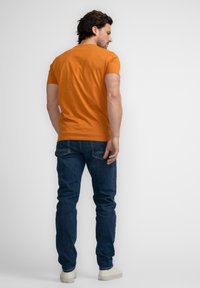 Camisa de manga corta color naranja, de material de algodón, con diseño ajustado. Combinada con jeans oscuros y zapatillas blancas, vista desde la espalda.