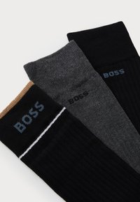 Tre par ribbade strumpor i svart, mörkgrå och en ljusare grå, med en topp i tanrand och "BOSS"-logotyp på varje.