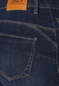 Jeans de mezclilla color azul oscuro con una etiqueta de cuero marcada "ONLY". Presenta bolsillos traseros estándar y costuras amarillas en contraste.