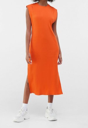 Geribbelde, oranje jurk met een ronde halslijn en mouwloos ontwerp. Accentuering met een zijslit. Gecombineerd met witte sneakers en enkelkousen.