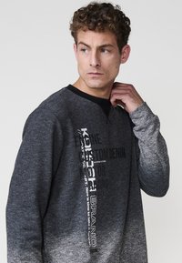 Felpa grigia scura con un design a gradiente, colletto a girocollo nero, maniche lunghe e grafica testuale prominente in bianco e nero.