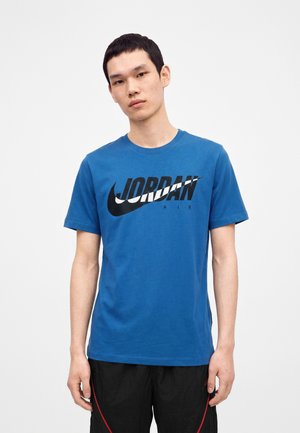 Jeune homme portant un t-shirt bleu Jordan Air et un pantalon noir, debout devant un fond clair uni.
