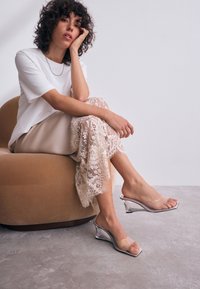 Prozirne sandale na platformu s transparentnim remenom preko prstiju. Dizajn uključuje metalik srebrni detalj, u kombinaciji s bež suknjom od čipke.