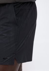Shorts de sport noirs avec poches latérales portés par une personne, avec un petit logo Nike près de l'ourlet sur la jambe gauche.