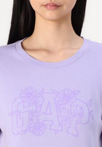 Levandulové bavlněné tričko s krátkým rukávem, s jemným fialovým květinovým vzorem kolem slova "GAP" na přední straně.