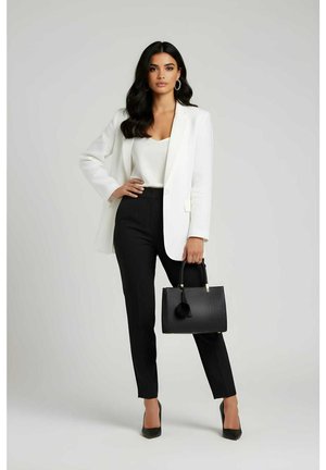 Blazer blanco, pantalones ajustados negros y un bolso negro con una borla. Modelo con tacones altos, de pie con la mano en la cadera, cabello corto y ondulado.