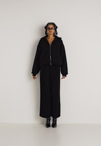 Crna zip-up oversized dukserica s spuštenim ramenima, uparena s labavim crnim hlačama i sjajnim crnim gležnjačama. Doplata su sunčane naočale.