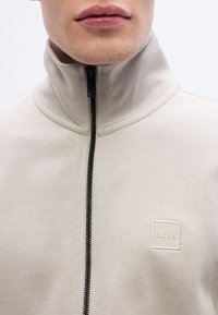 Ανοιχτό μπεζ μπουφάν με φερμουάρ και ψηλό γιακά, υφασμα με υφή, μαύρο φερμουάρ και μικρό τετράγωνο λογότυπο στην αριστερή πλευρά του στήθ ους.