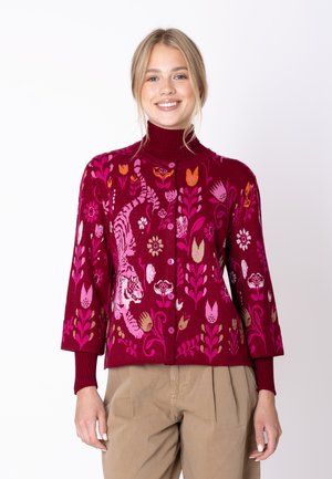 Giovane donna sorridente, indossa un maglione bordeaux con bottoni con motivi floreali e di tigre rosa e arancioni, abbinato a pantaloni beige.