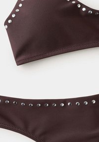 Haut et bas de bikini marron foncé avec des clous ronds argentés le long des bords, posés à plat sur une surface blanche.