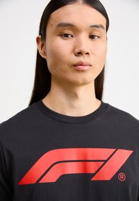 Black cotton T-shirt featuring a prominent red Formula 1 logo on the front. Crew neck and short sleeves, smooth texture.