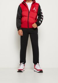 Czerwona pikowana kamizelka z czarnymi rękawami, zamkiem błyskawicznym i logo. W połączeniu z czarnymi spodniami dresowymi i czerwono-białymi butami sportowymi.