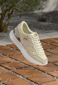 Μπεζ αθλητικό παπούτσι με διακριτικά γεωμετρικά μοτίβα, απαλό σουέντ και υφή στο πλάι. Διαθέτει λευκή σόλα και μεταλλικές χρυσές λεπτομέρειες.
