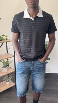 Zwarte polo met witte kraag en dunne horizontale strepen, gecombineerd met blauwe denim shorts. De achtergrond bevat groene planten en een plankeenheid.