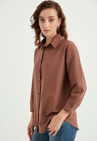 Camicia marrone con bottoni, colletto classico e maniche lunghe, realizzata in tessuto liscio. Indossata con jeans blu, caratterizzati da una vestibilità comoda.