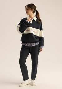 Granatowy i kremowy sweter typu color-block z haftowanym logo, nałożony na paskowaną koszulę, w połączeniu z czarnymi spodniami i białymi trampkami.