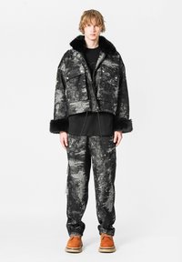 Chaqueta de abrigo de patrones en negro y gris con cuello de piel sintética, combinada con pantalones a juego. El modelo lleva calzado marrón.