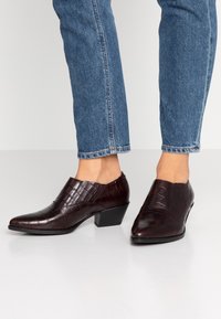 Bruna ankelskor med krokodilmönster, bärs med blå jeans, visar en låg klack och en slank design mot en neutral bakgrund.