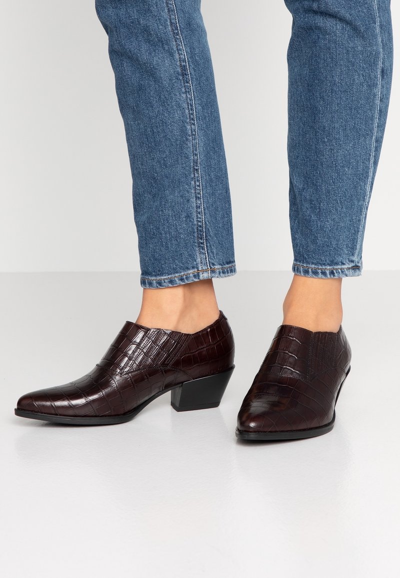 Bruna ankelskor med krokodilmönster, bärs med blå jeans, visar en låg klack och en slank design mot en neutral bakgrund.