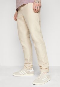 Beige slim-fit broek gemaakt van lichtgewicht stof, met een zachte textuur. G gedragen met grijze sneakers, met witte strepen en veters.
