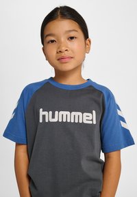 Grå t-shirt med blå raglanärmar pryds av en vit "hummel"-logotyp framtill. Mjukt tyg, avslappnad passform med ränder på ärmarna.