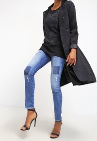 Cappotto lungo in finta pelle scura su una maglietta nera con design metallico, abbinato a jeans skinny blu scuro strappati con dettagli di toppe e tacchi neri.