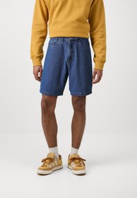 Μπλε τζιν σόρτς σε ανοιχτό χρώμα με ευθεία γραμμή, χαρακτηριστικά κουμπιοθήκη και δύο μπροστινές τσέπες. Συνδυασμένα με ένα κίτρινο crewneck φούτερ και αθλητικά παπούτσια.