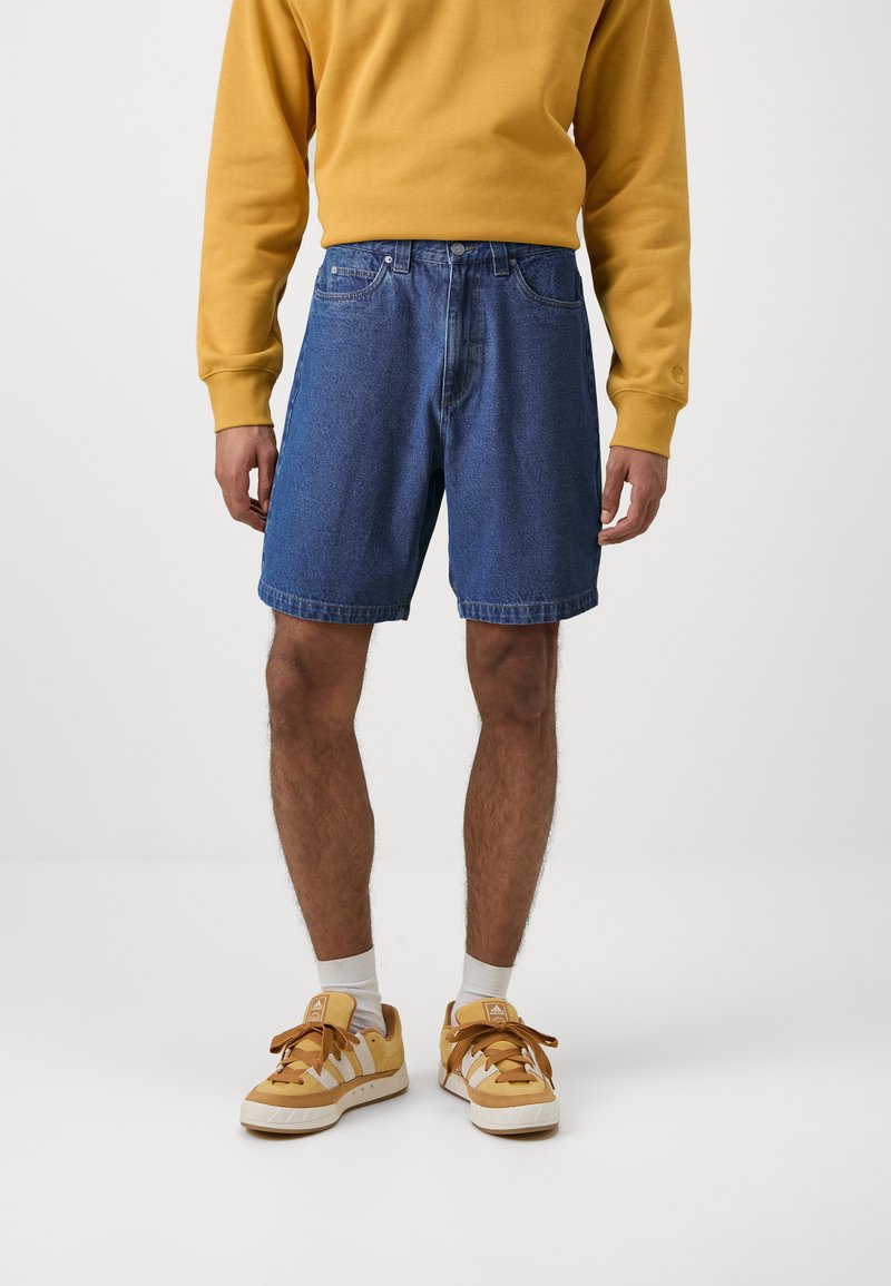 Μπλε τζιν σόρτς σε ανοιχτό χρώμα με ευθεία γραμμή, χαρακτηριστικά κουμπιοθήκη και δύο μπροστινές τσέπες. Συνδυασμένα με ένα κίτρινο crewneck φούτερ και αθλητικά παπούτσια.