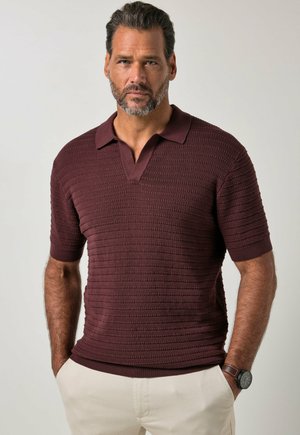Mann mit Bart, der ein strukturiertes dunkelrotes kurzärmeliges Poloshirt und beige Hose trägt, steht mit den Händen in den Taschen vor einem hellen Hintergrund.