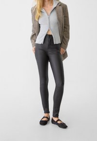 Pantalon noir taille haute avec une finition brillante, associé à un haut court gris clair zippé et un blazer marron à carreaux. Chaussures plates noires.
