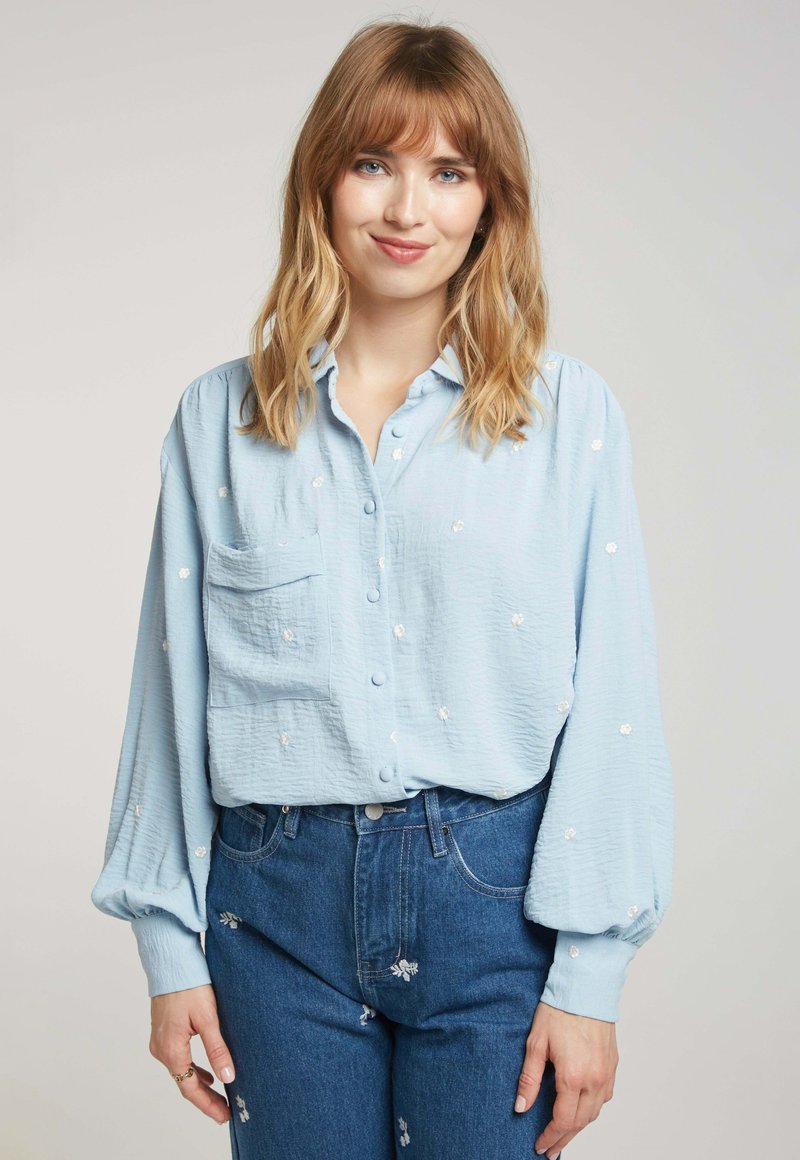 Chemisier bleu clair, à boutons, en tissu texturé, présentant une poche avant et des broderies florales blanches, associé à un jean bleu taille haute.